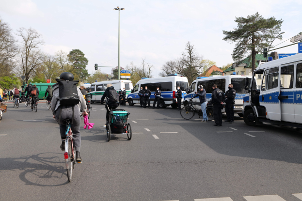 Gruppe von Menschen auf Fahrrädern auf einer Straße mit Polizeiwagen, anderen Fahrzeugen, Bäumen, Laternen, Verkehrsampeln, Schildern, Gebäuden und einem bewölkten Himmel.