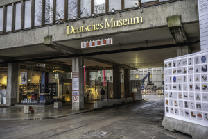 Das Deutsche Museum in Berlin, Deutschland, ein großes Gebäude mit Glasfenstern, Säulen und einer Namensplakette, mit einer Tafel mit Bildern und Text auf der rechten Seite und anderen Gebäuden, Bäumen und dem Himmel im Hintergrund.