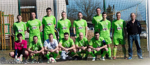 Eine Gruppe von Männern in grünen T-Shirts steht und hockt auf einem Fußballfeld mit einem Ball davor, ein Torpfosten rechts und ein Hintergrund aus Bäumen, Gebäuden, Pfosten, Fahrzeugen und dem Himmel, mit Text, der Zahlen am unteren linken Rand auflistet.