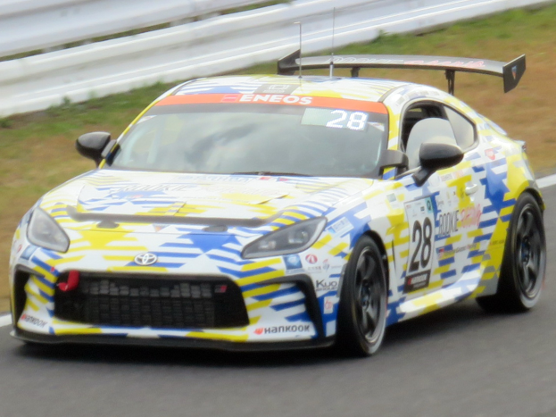 Ein weißer Toyota Supra GT3-Rennwagen mit Sponsorentext und Nummern auf einer Rennstrecke, umgeben von einer Metallschranke und Gras.