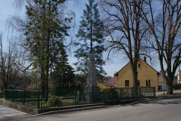 Denkmal für Holocaust-Opfer in der Mitte einer Straße, umgeben von einem Metallzaun, Grünfläche, Häusern, einem Straßenschild und einem bewölkten Himmel.