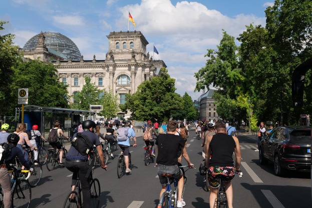 Eine Gruppe von Menschen, die auf Fahrrädern eine von Bäumen gesäumte Straße in Berlin, Deutschland, entlangfahren, mit Gebäuden und einer Bushaltestelle im Hintergrund, unter einem bewölkten Himmel und einer Flagge auf einem Gebäude.