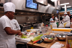 Eine Gruppe von Köchen in einer Küche bei der Zubereitung von Essen auf einem Schneidbrett, mit einem Koch im Vordergrund in einer weißen Schürze und einer weißen Haube, der ein Messer hält und von Utensilien, Schüsseln, Gemüse, einem Handy, Flaschen und anderen Gegenständen umgeben ist, mit einem Fernseher und Deckenlampen im Hintergrund.