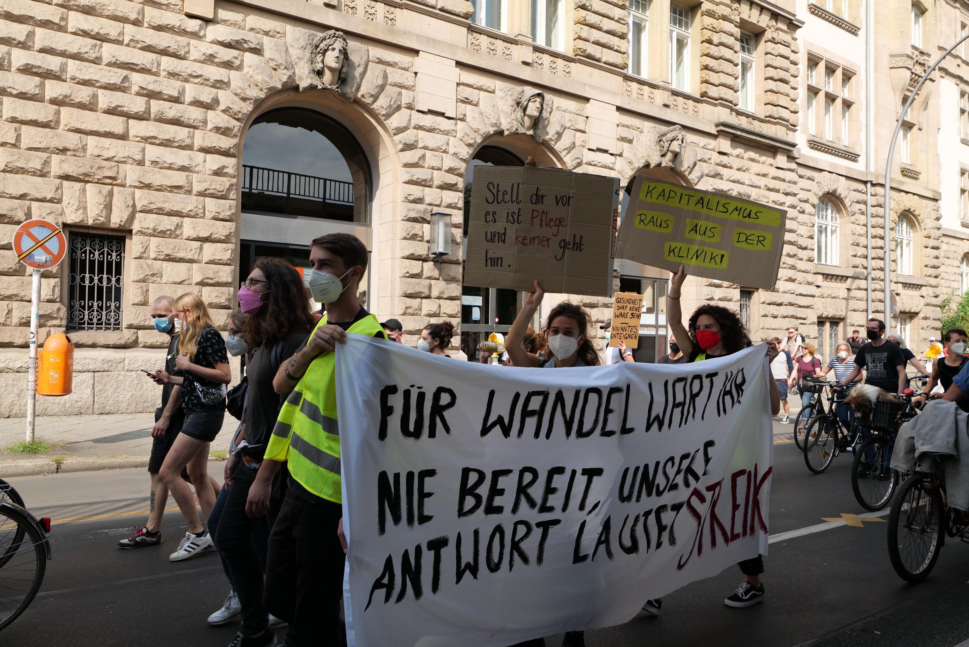 Eine Gruppe von Menschen marschiert auf einer Stadtstraße in einem Protest, einige halten Schilder und andere fahren Fahrräder, mit einem dekorierten Gebäude, Bäumen und einem Laternenpfahl im Hintergrund.