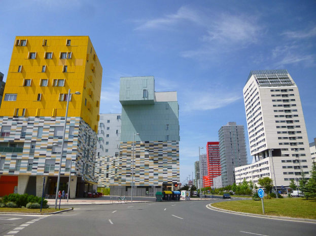 Eine Straße in einer Stadt mit hohen Gebäuden im Hintergrund, Straßenmäste, Straßenlaternen, Kraftfahrzeuge, Infotafeln, Sträucher, Bäume und ein bewölkter Himmel, mit dem einzigartigen Architekturstil der Universität Rotterdam.