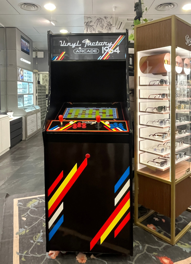 Ein Geschäft mit einem alten Arcade-Automaten in der Mitte, einem Glasvitrine auf der rechten Seite, Schränken und einem Fernseher im Hintergrund und einer Deckenbeleuchtung, die eine retro-mäßige Atmosphäre schafft.