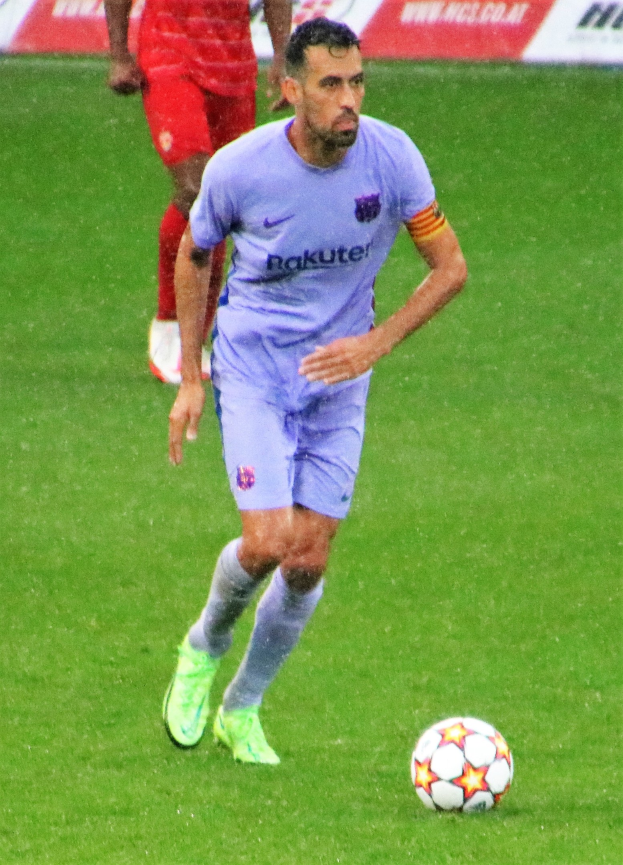 Fussballspieler in Sportbekleidung und -schuhen auf einem Feld mit einer Texttafel im Hintergrund.