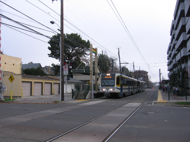 Ein Lichtschnellbahnzug fährt eine Straße mit hohen Gebäuden, Strommasten mit Drähten, einigen Fußgängern, Bäumen und einem klaren blauen Himmel im Hintergrund entlang.