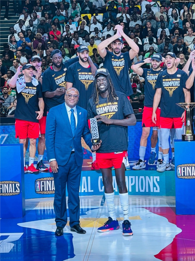 Männer mit Trophäen auf einem Basketballfeld mit Zuschauern im Hintergrund und zwei Trophäen auf Podesten.