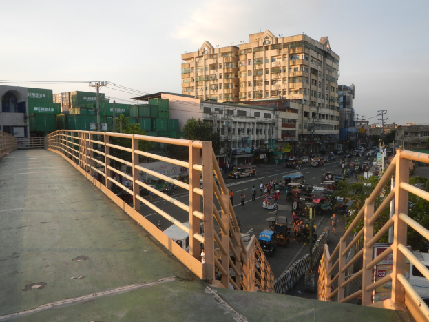 Ein Fußgängerbrücke mit Geländern überspannt eine vielbefahrene Straße mit Fahrzeugen und Fußgängern, im Hintergrund sind Gebäude, Bäume, Strommasten und ein bewölkter Himmel zu sehen.