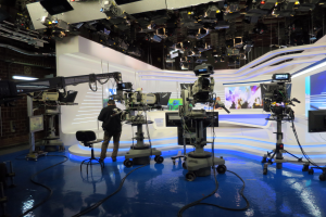 Ein Mann steht vor einem Fernseher und hält ein Mikrofon, umgeben von Kameras, einem Stuhl, einem Bildschirm, einer Wand und einer Decke mit Lampen, was auf eine Live-Übertragung hindeutet.