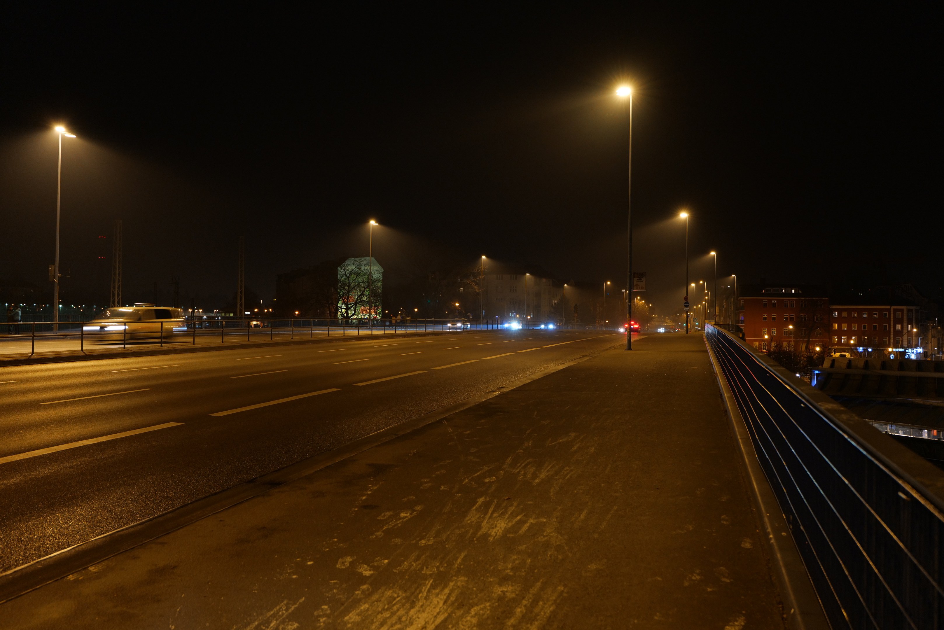 Langzeitbelichtung einer nächtlichen Stadtstraße mit fahrenden Fahrzeugen, Geländern, Laternen, Gebäuden, Bäumen und einem dunklen Himmel.
