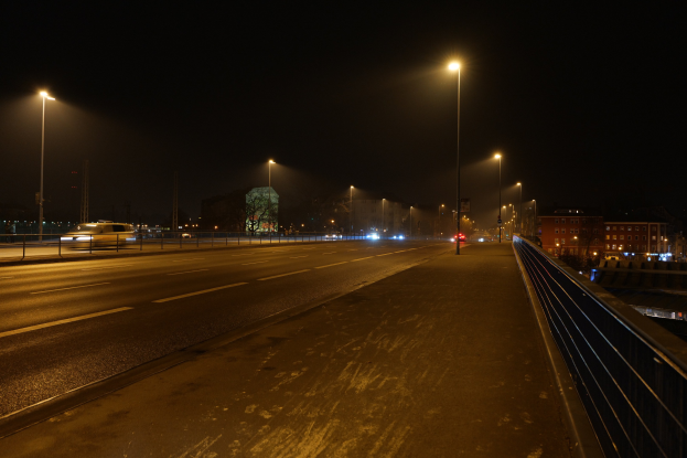 Langzeitbelichtung einer nächtlichen Stadtstraße mit fahrenden Fahrzeugen, Geländern, Laternen, Gebäuden, Bäumen und einem dunklen Himmel.