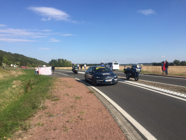 Eine Gruppe von Menschen steht neben einem Polizeiauto und einem Motorrad auf der Seite einer Straße, mit einer Fahne und einem bewölkten Himmel im Hintergrund.