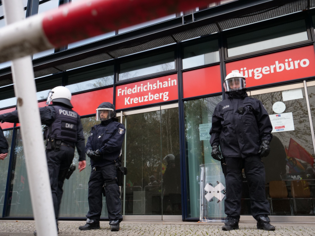 Polizeibeamte in Uniform vor einem gläsernen Gebäude mit einer Stange links und Schildern mit "Friedrichshain Kreuzberg"-Text im Hintergrund.