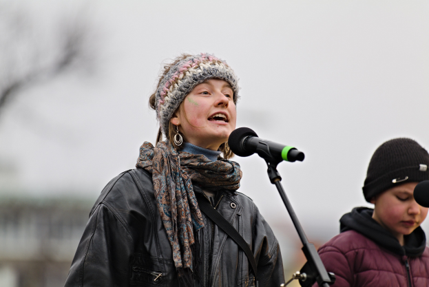 Eine Frau in schwarzer Jacke, Schal und Mütze singt in ein Mikrofon auf der Bühne, neben ihr eine Person in einer maronenfarbenen Jacke und schwarzer Mütze, während das Publikum und der Hintergrund unscharf sind und einen traumhaften Effekt erzeugen.