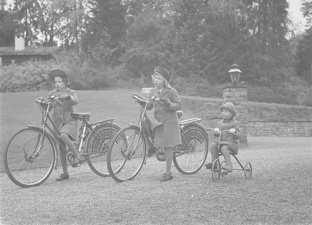 Drei Kinder fahren Fahrräder in einem Park, tragen Mäntel und Mützen, wobei eines auf einem Dreirad sitzt; Bäume, Pflanzen, ein Haus und ein Laternenpfahl sind im Hintergrund zu sehen.