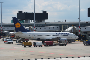 Ein Lufthansa Airbus A320-200 steht auf dem Frankfurter Flughafen geparkt mit Fahrzeugen und Wagen auf der Straße davor, einem Gebäude, Laternenpfählen und einem klaren blauen Himmel im Hintergrund.