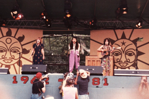Drei Mädchen auf der Bühne, zwei singen in Mikrofone und eines spielt Gitarre, mit Publikum, das Fotos vor der Bühne macht und Bühnenausrüstung im Hintergrund.