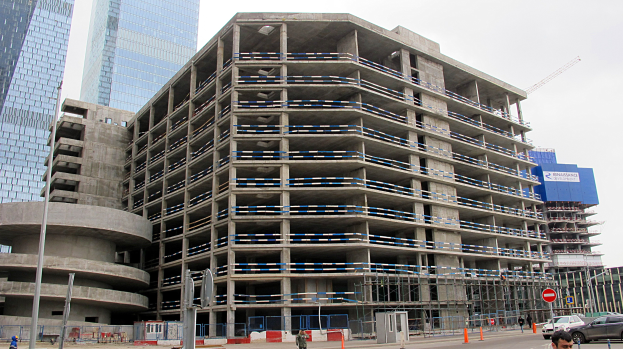Großes Bauprojekt in einer Stadt umgeben von Wolkenkratzern, Fahrzeugen, Fußgängern, Verkehrsinfrastruktur und Kränen unter einem klaren Himmel.