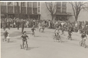 Eine Gruppe von Kindern auf Fahrrädern, die eine Straße entlangfahren, mit einer Menge von Menschen am Straßenrand, Bäumen und Gebäuden im Hintergrund und Text am unteren Rand des Bildes, alles in Schwarz-Weiß.