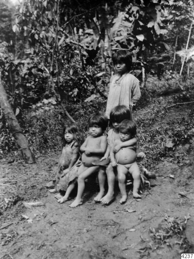 Fünf Kinder sitzen auf einer Schotterstraße in einem Schwarz-Weiß-Bild mit Bäumen und einer stehenden Person im Hintergrund und Text unten; die Kinder scheinen beunruhigt zu sein.