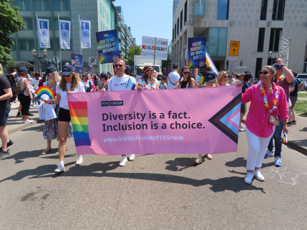 Eine Gruppe von Menschen marschiert auf einer Straße, hält ein Banner mit der Aufschrift "Diversität ist eine Tatsache, Inklusion ist eine Wahl" in den Händen, einige tragen Mützen und Schutzbrillen, andere zusätzliche Banner, vor einem Hintergrund aus Gebäuden, Bäumen und einem klaren blauen Himmel.
