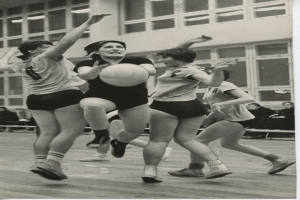 Eine Gruppe von Frauen beim Volleyballspielen auf einem Platz mit einem Ball in der Mitte, Menschen auf Bänken sitzend und ein Gebäude mit Fenstern im Hintergrund sowie Text am unteren Bildrand.