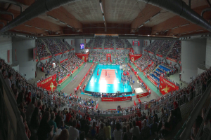 Eine große Halle voller Zuschauer bei einem Volleyballspiel mit Bannern, Bildschirmen, Deckenlampen und Geländern unten.