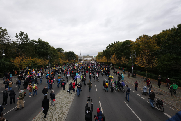 Eine große Gruppe von Menschen, die eine von Bäumen gesäumte Straße mit Laternenpfählen entlanggeht und während einer Protestmarsch in Berlin Kameras hält, mit einem Gebäude und einem klaren Himmel im Hintergrund.