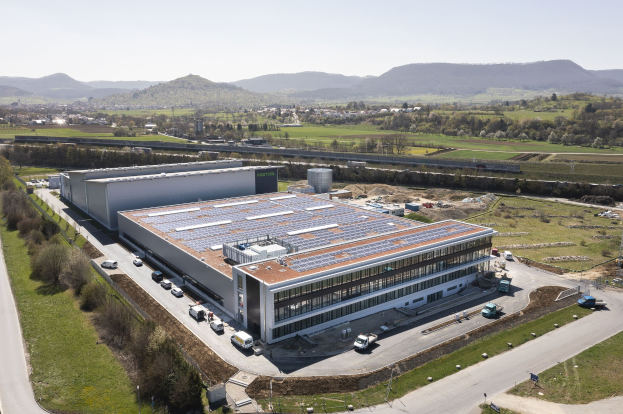 Luftaufnahme eines großen Gebäudes mit Solarpaneelen auf dem Dach, umgeben von Fahrzeugen, Bäumen, Gras und Masten, vor Bergen und einem klaren blauen Himmel, als Deutschlands erste solarbetriebene Fabrik identifiziert.