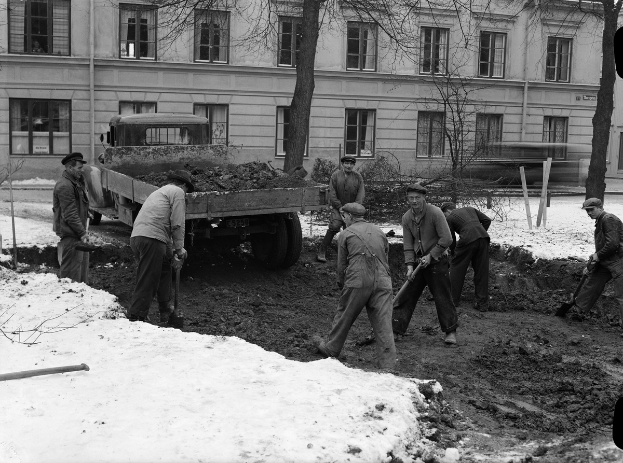 Eine Gruppe von Männern in Mützen schippt Schnee auf einer Baustelle, mit einem Lastwagen im Hintergrund, umgeben von Bäumen und Gebäuden mit Fenstern, dargestellt in Schwarz-Weiß.
