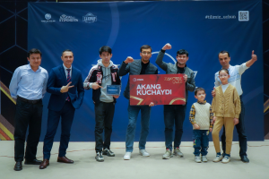 Eine Gruppe von Menschen auf einer Bühne, die eine Tafel mit dem Text "Akang Kuchaydi - Zakovat Quiz" halten, mit einem Banner, Lichtern und anderen Gegenständen im Hintergrund.