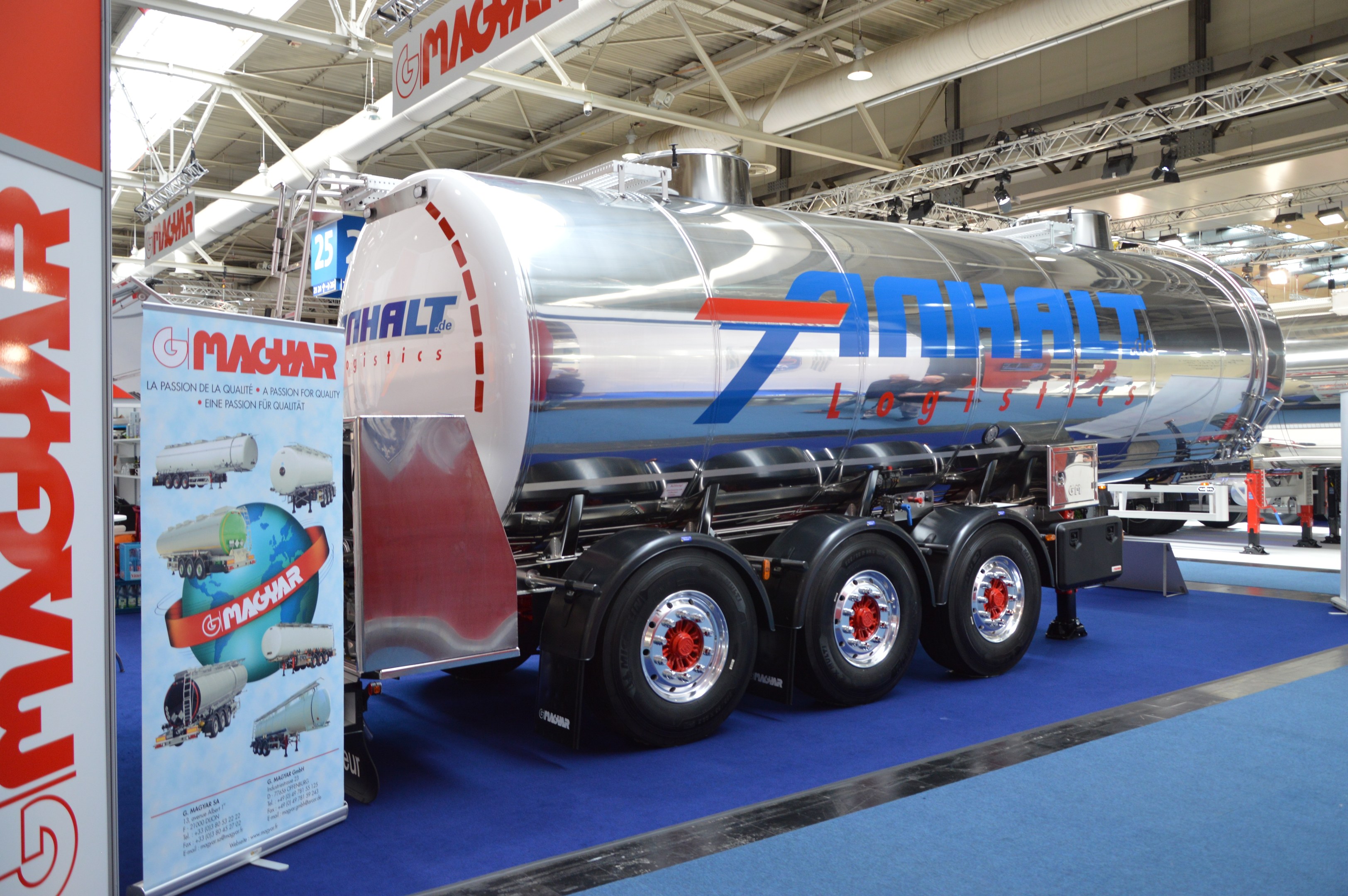 Großer Tanklastzug auf blauen Teppichboden bei einer Messe ausgestellt, umgeben von Bannern, Lichtern, Stangen und anderen Fahrzeugen.