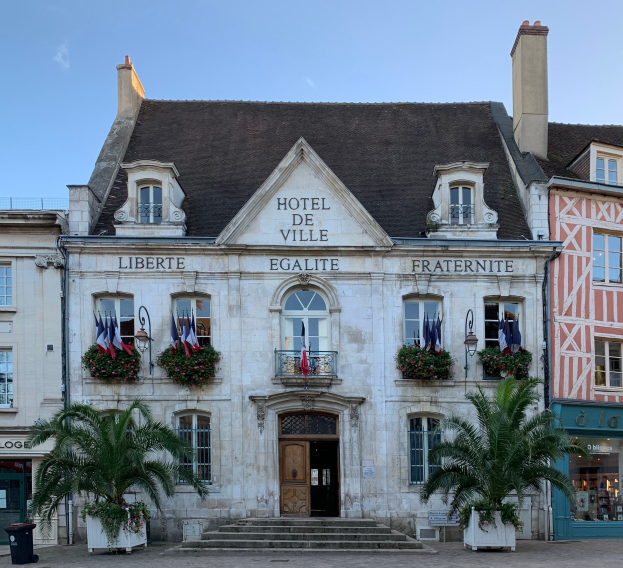 Das Hotel de Ville in Saint-Emilio, Frankreich, ein Gebäude mit Fenstern, Türen und Text an der Wand, umgeben von Pflanzen mit Blumen, Bäumen, einem Müllcontainer und einer Treppe unter einem bewölkten Himmel.