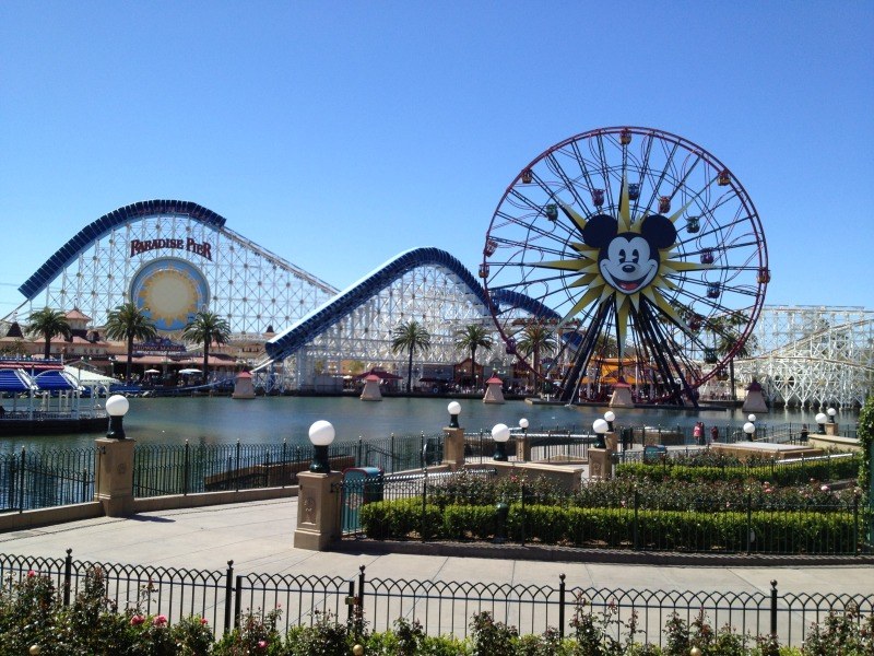Disneylands California Adventure Vergnügungspark mit einem Riesenrad, Achterbahn, Grünflächen, Laternenmasten, einem Wasserbecken im Vordergrund und einem hellblauen Himmel.
