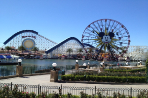 Disneylands California Adventure Vergnügungspark mit einem Riesenrad, Achterbahn, Grünflächen, Laternenmasten, einem Wasserbecken im Vordergrund und einem hellblauen Himmel.