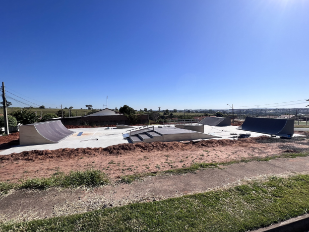 Skate-Anlage mit mehreren Rampen, Grünfläche, Bäumen, Pfählen, Drähten und einem klaren blauen Himmel.