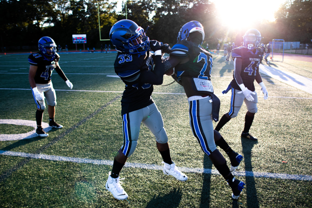 Gruppe junger Männer in Helmen und Schuhen, die Fußball auf einem Feld mit Bäumen und einem klaren blauen Himmel spielen.