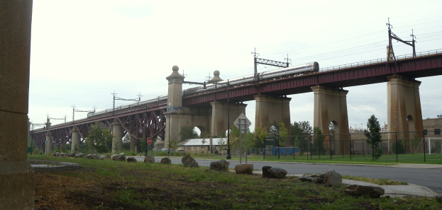 Eine Bahn überquert eine Brücke mit Säulen, Strommasten, Schildern, einem Zaun, Gras, Steinen, einer Straße, Bäumen, Gebäuden und einem bewölkten Himmel im Hintergrund.