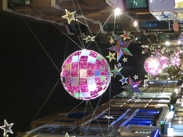 Eine nächtliche Straßenansicht einer Stadt, die mit Lichtern und Dekorationen gefüllt ist, mit einer großen Diskokugel, die von einem Gebäude hängt, und beleuchteten Gebäuden im Hintergrund.