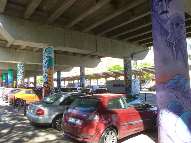 Ein Parkplatz mit zahlreichen geparkten Autos unter einer Brücke, mit Graffiti an den Pfeilern und nächsten Gebäuden, mit Bäumen im Hintergrund und einem Van unter den Fahrzeugen geparkt.