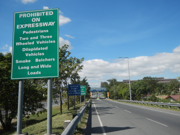 Grünes Autobahnschild mit der Aufschrift "Verboten auf der Autobahn" mit Fahrzeugen, Laternenpfählen, Bäumen, Gebäuden und bewölktem Himmel im Hintergrund.
