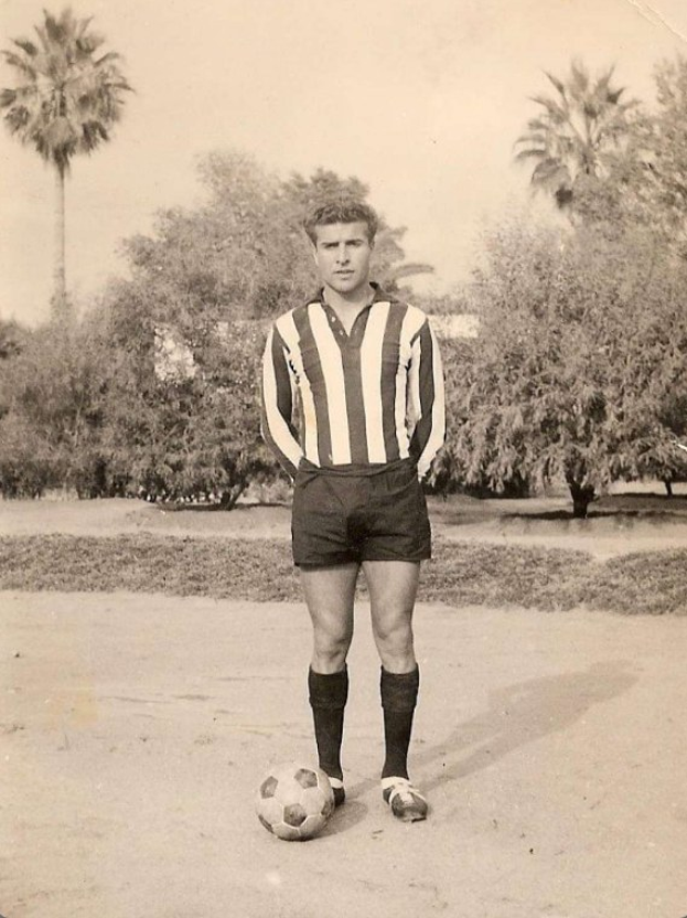 Schwarzes und weißes Foto eines älteren Mannes, der einen Fußball auf einem Rasen hält.