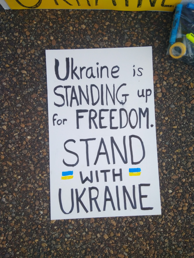 White paper with "Ukraine is Standing Up for Freedom, Stand with Ukraine" text on the ground in front of a yellow banner, with a plastic cover containing various objects to the right.