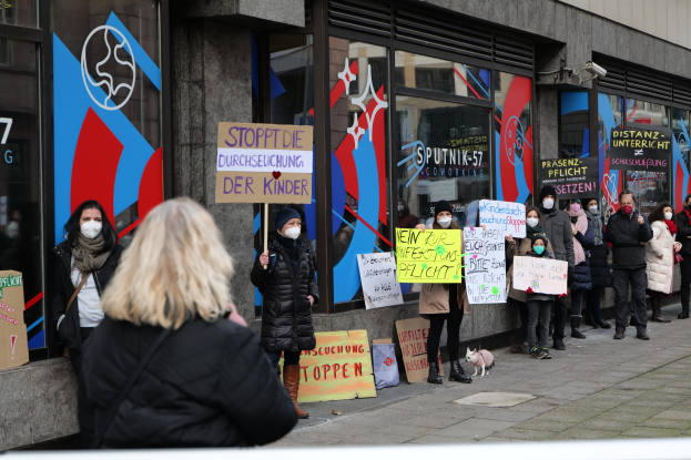 Gruppe von maskierten Menschen mit Schildern vor einem Glasfrontgebäude, mit Protesttafeln, Kameras und einem Hund.