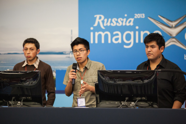 Drei Jungen stehen neben einem Schreibtisch mit zwei Computersystemen, einer spricht in ein Mikrofon und trägt eine ID-Karte mit roten Tags, vor einer Wand mit einem Werbeplakat, auf dem "Russia Imagine 2013" steht.