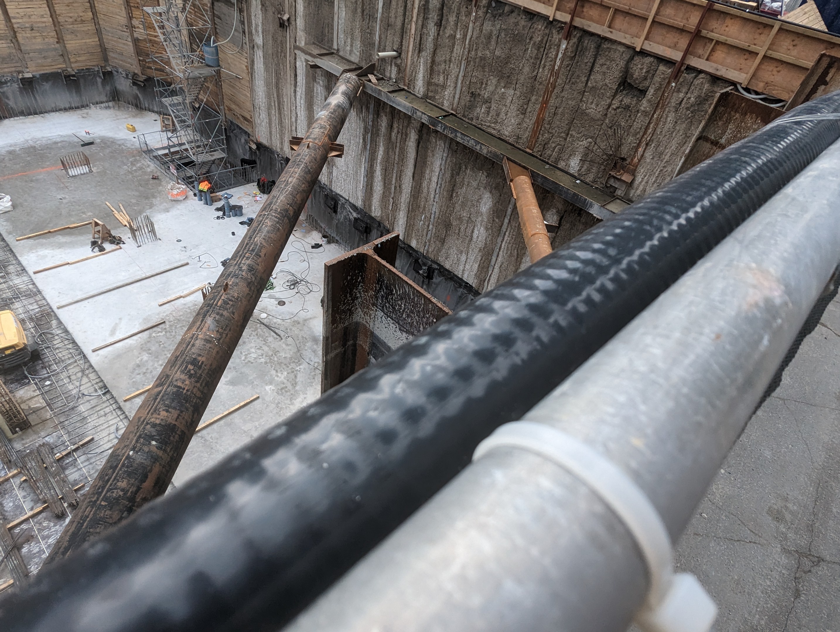 Großes Metallrohr auf einer Baustelle mit Arbeitern, Holzwerkstoffen und Geräten, mit einer Person in einem blauen Hemd auf der rechten Seite und Fahrzeugen und einem Gebäude im Hintergrund.