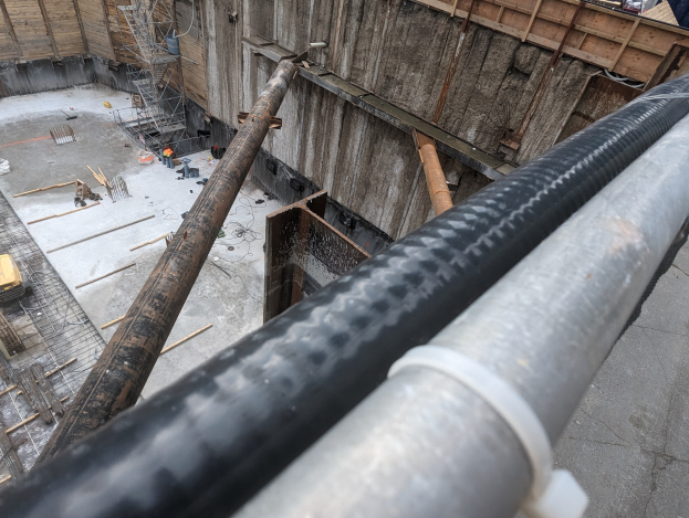 Großes Metallrohr auf einer Baustelle mit Arbeitern, Holzwerkstoffen und Geräten, mit einer Person in einem blauen Hemd auf der rechten Seite und Fahrzeugen und einem Gebäude im Hintergrund.