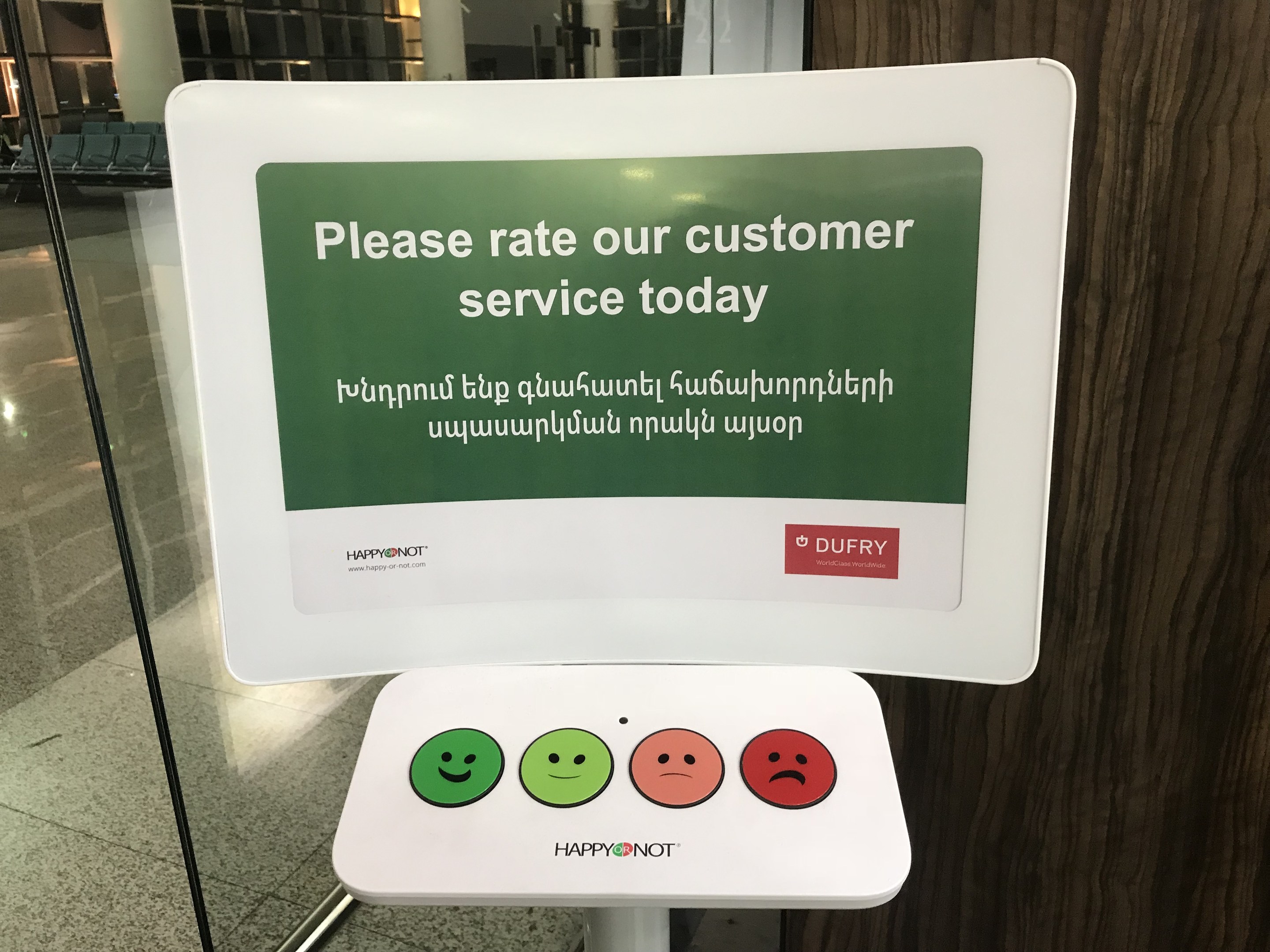 Ein Schild mit der Aufschrift "Bewerten Sie unseren Kundenservice heute" steht vor einer Glastür, mit einer hölzernen Wand rechts und einem Boden darunter, vor einem Hintergrund aus Stühlen, Säulen und Lampen.